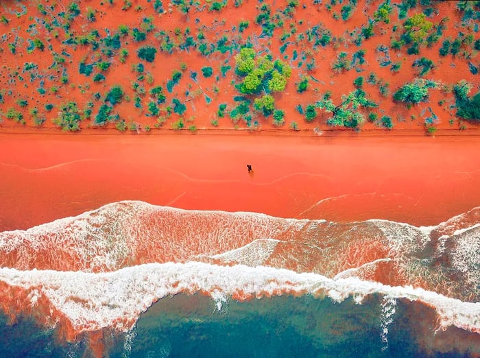 An aerial view captures a vibrant red sandy beach in Western Australia, with a solitary hiker wandering along its shore.