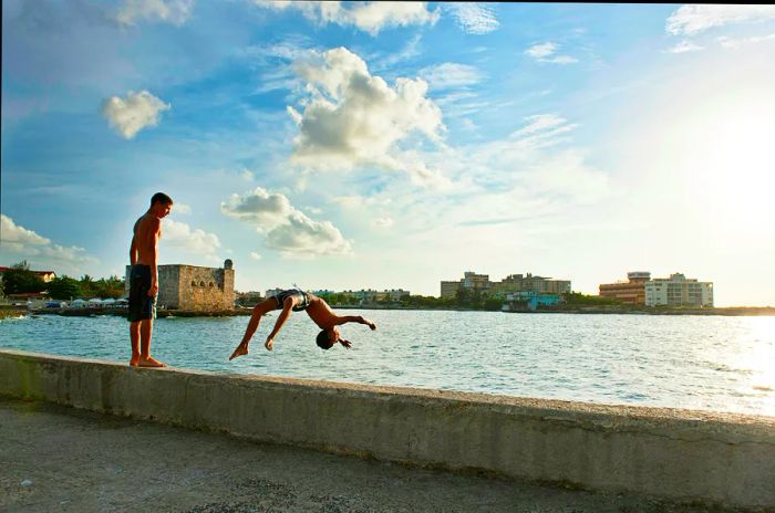 A boy watches from the harbor wall in Havana as another boy performs a backflip into the water below.