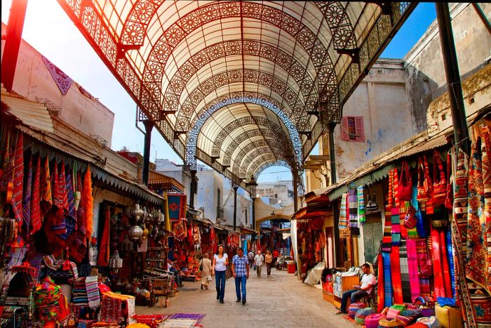 Shoppers explore a covered market filled with vibrant rugs and scarves.