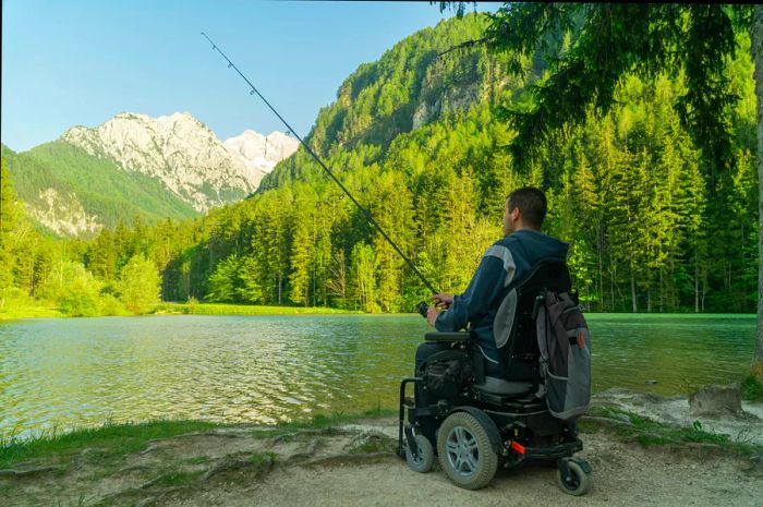 A young man in a wheelchair is fishing at a stunning lake on a sunny day, with mountains visible in the background.