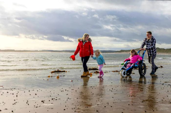 A family with two children enjoys the beach in Beadnell, North East England. The older girl, a wheelchair user, is utilizing a beach wheelchair.