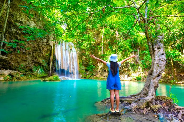 A teenager joyfully stands with arms raised in front of a waterfall cascading into a turquoise pool, with her back to the camera.