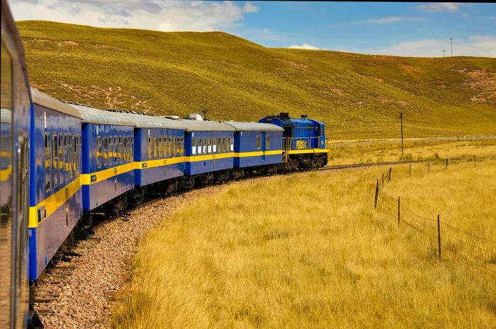 A vibrant blue-and-yellow train curves around a bend in Peru's lush landscape.