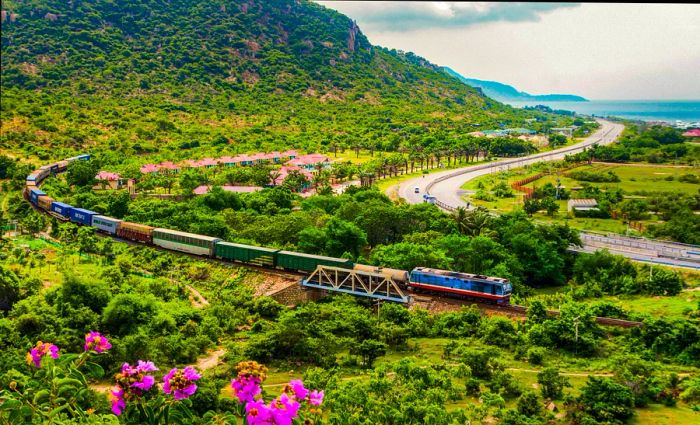 A train glides through a vibrant landscape, flanked by lush trees and the shimmering coastline.