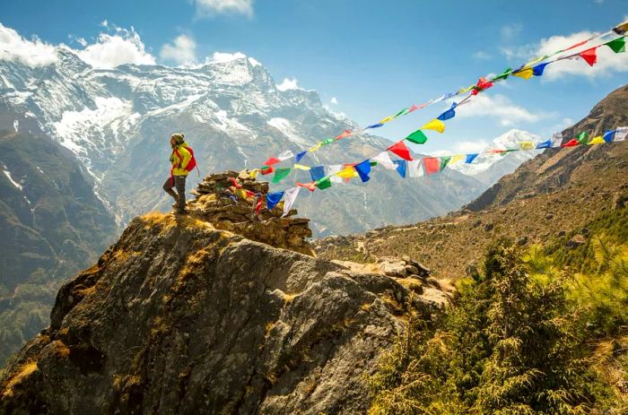 A hiker stands atop a towering mountain in Nepal, with colorful prayer flags fluttering in the breeze.