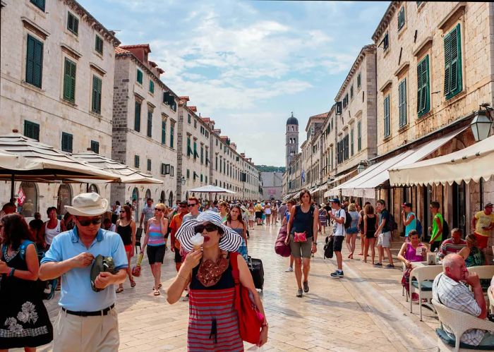 A lively summer scene on the main street (Stradun or Placa), featuring locals, tourists, and a woman enjoying an ice cream cone.