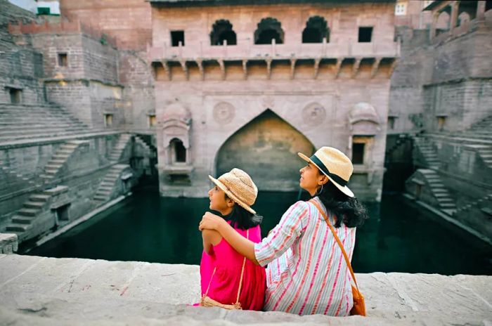 A mother and daughter, seen from behind, relax at the Toorji Ka Jhalra stepwell in Jodhpur, Rajasthan, while wearing hats.