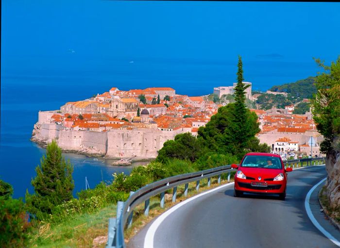 A red car winding around a scenic road with the red rooftops of Dubrovnik visible in the distance