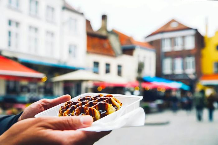 A person enjoys a takeaway tray filled with a waffle drizzled in chocolate sauce in a medieval square.