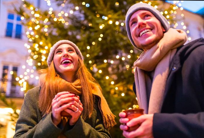 A couple dressed in warm winter attire enjoying hot beverages with a beautifully lit Christmas tree behind them.