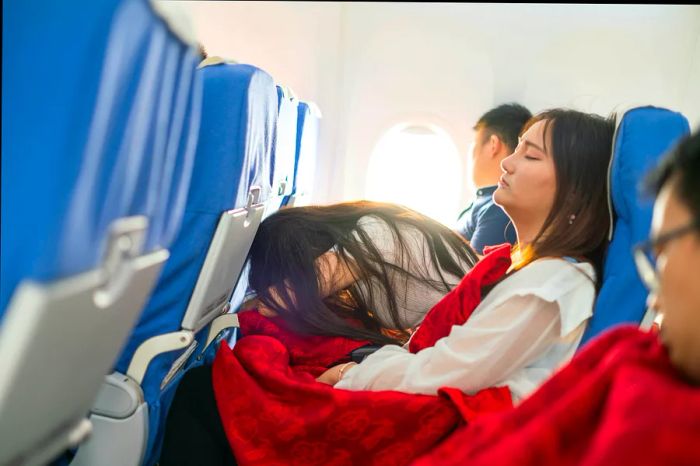 In an airplane cabin, a passenger in the middle seat is leaning over and dozing off on the tray table.