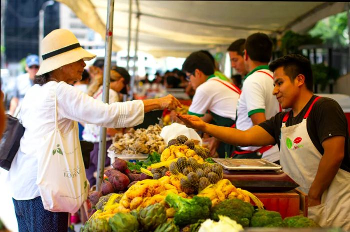 A woman in a white shirt and hat exchanges money with a vegetable vendor at a market in Miraflores, Lima, Peru