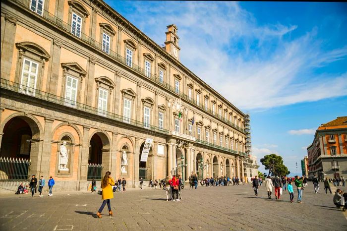 Tourists stroll in front of the grand facade of the historic Royal Palace in Naples.