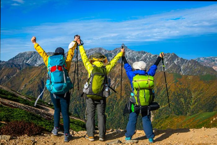 Three hikers stand triumphantly with their arms raised, gazing out towards a stunning mountain range.