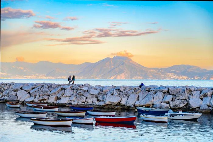 Two individuals stroll along the Napoli waterfront at twilight.