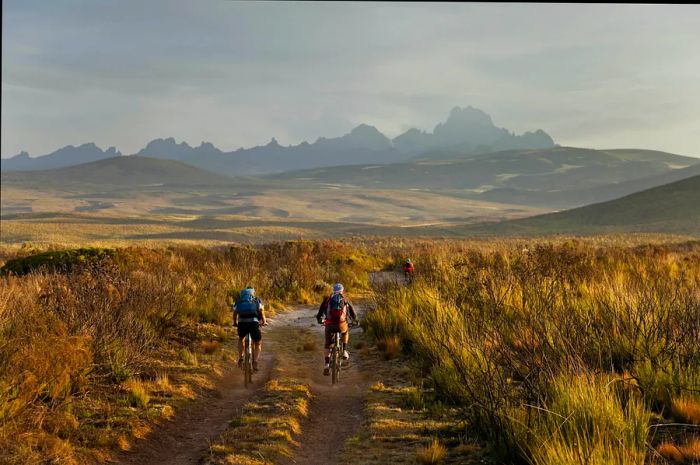 Mountain bikers ride along a dirt trail toward a backdrop of majestic peaks