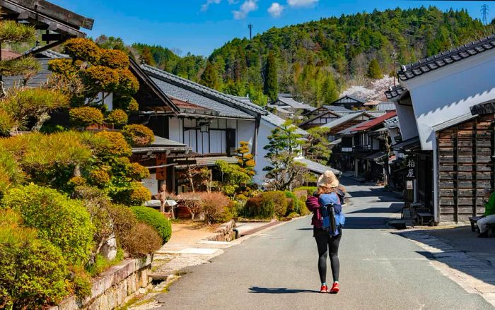 A lone hiker traverses the Nakasendō path, passing through a quaint traditional Japanese village with wooden low-rise homes lining the way.