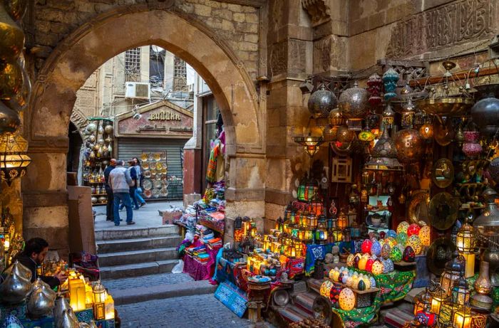 A vibrant stall filled with lamps and lanterns in a souk, with a pathway leading to more stalls through an arched entrance.