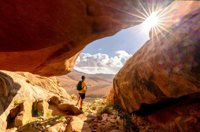 A person stands inside a sandstone rock cave, watching the sunset in Barranco de las Penitas, Fuerteventura, Canary Islands, Spain