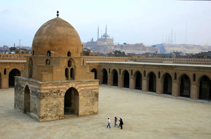 Four individuals stroll through a courtyard adorned with a grand domed structure and arched cloisters.