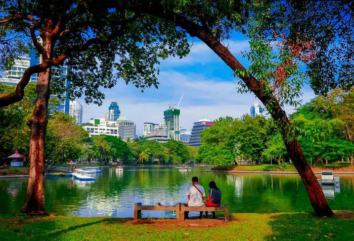 A couple relaxes on a bench overlooking the tranquil lake at Lumpini Park in Bangkok, Thailand.