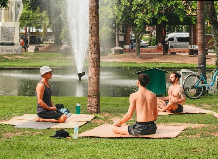 Three individuals practice yoga on the grass in a Chiang Mai park, with a small lake and fountain visible in the background.