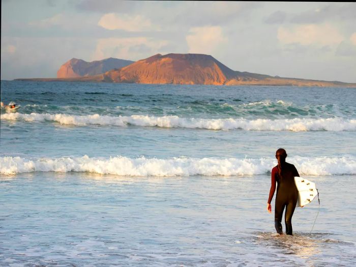 A surfer wades into the waves at Famara beach, with the stunning La Graciosa Island in the backdrop, Lanzarote, Canary Islands, Spain