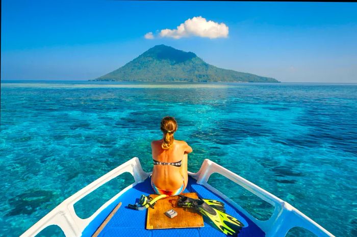 A young woman in a swimsuit lounges on a boat, equipped with a mask and flippers, gazing at the clear sea and the majestic Manado Tua volcano in North Sulawesi, Indonesia.