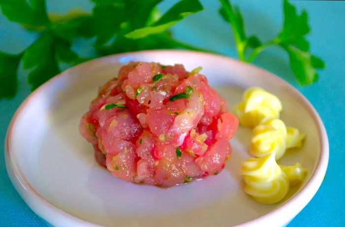 On a round white saucer, a raw palamita fish meatball, pink in color with small bits of chopped green parsley, sits beside three tufts of yellow mayonnaise. At the top, a tuft of green parsley is placed, against a turquoise tablecloth.