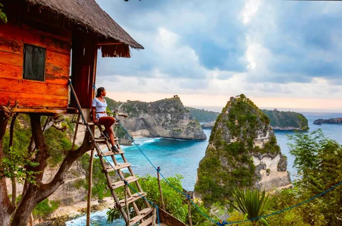 A solo woman perches atop a ladder leading to a treehouse that overlooks a bay dotted with rocky islets.