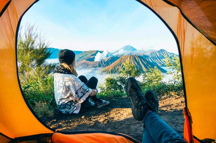 A woman sits with her back to a tent, gazing over a volcanic landscape