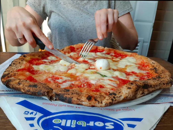 A person sits at a table with a large pizza on it in a blue-and-white box