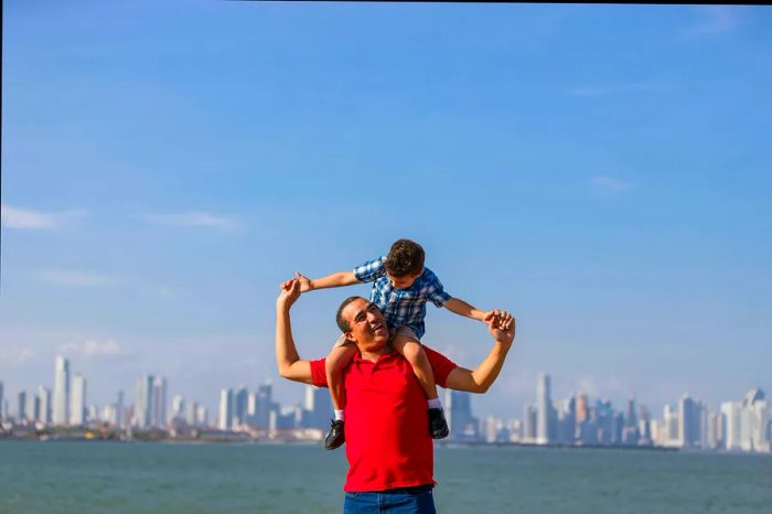 A young boy rides on a man's shoulders at the waterfront, framed by the skyline of Panama City.