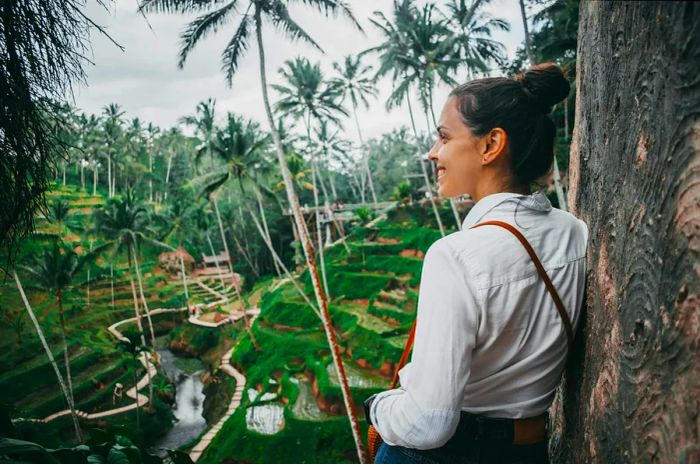 A young woman with brown hair in a white shirt gazes over the picturesque rice terraces and palm trees of Ubud, Bali.