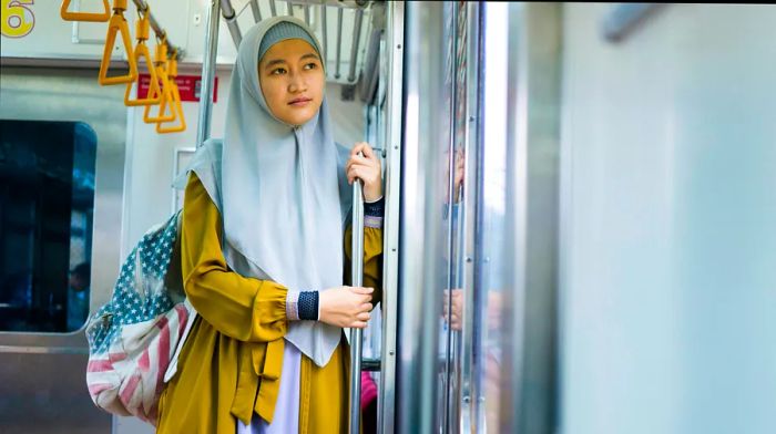 A young Muslim woman in a hijab and backpack stands at the entrance of a train in Jakarta, Indonesia.