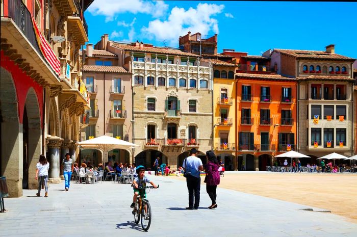 A child rides a bike while people stroll around Plaça Mayor in Vic, Spain