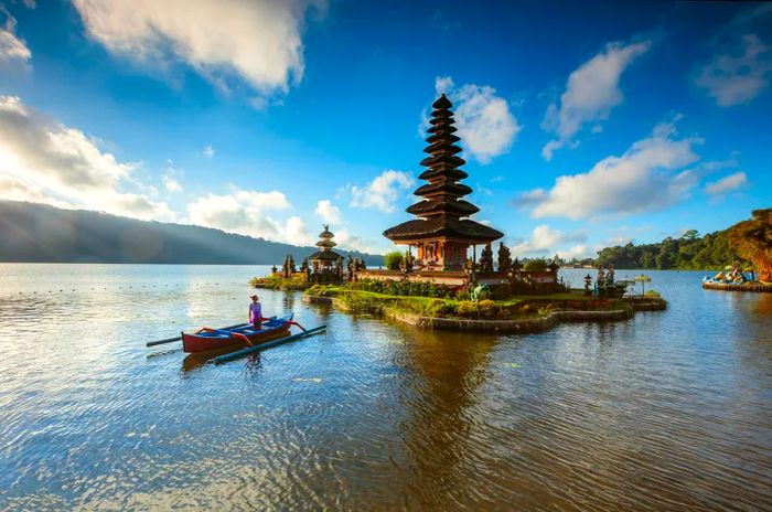 A Balinese woman paddles her boat towards the iconic Ulun Danu Beratan Temple in Bedugul, Bali, framed by clear blue skies and fluffy clouds.