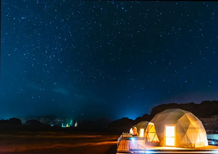 A series of dome-shaped tents set against the desert landscape of Wadi Rum, illuminated by a starry night sky.