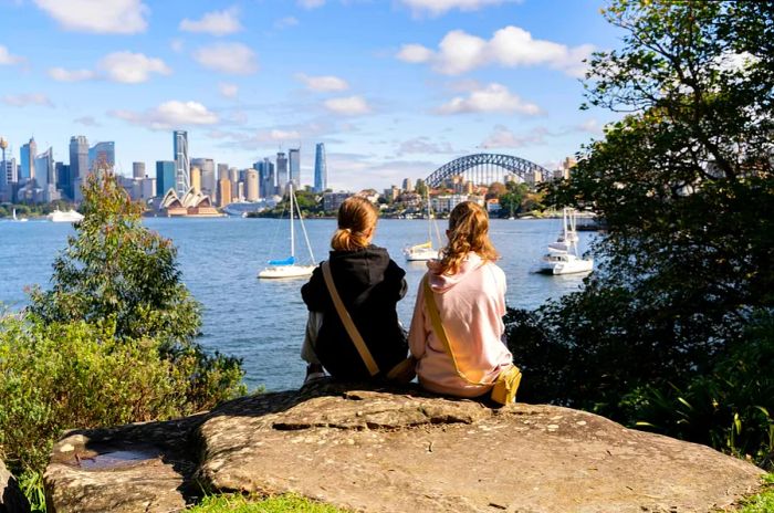 Two girls perched on a rock, gazing out at an iconic harbor featuring a white opera house and a grand bridge.