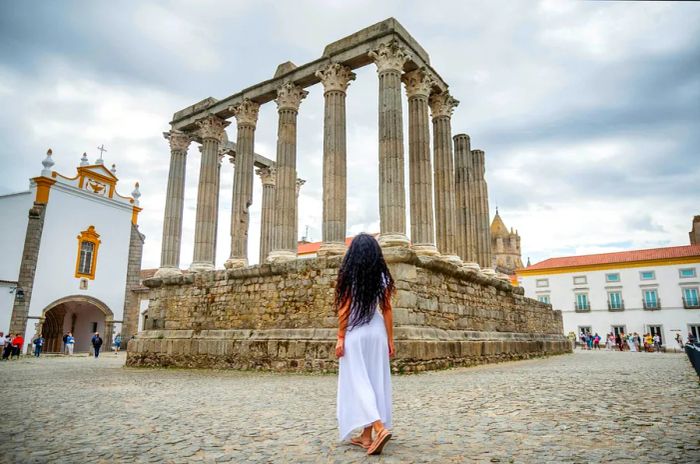 A rear view of a Portuguese woman in a flowing white dress standing before the Roman ruins of the Temple of Diana in Évora, Portugal.