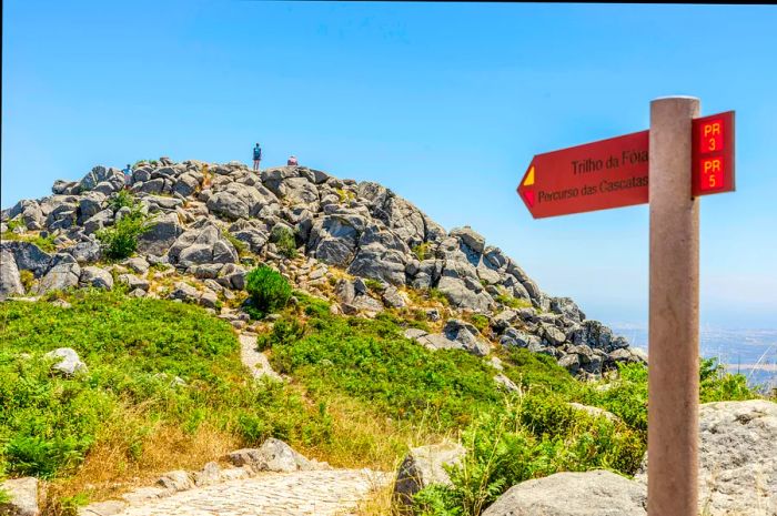 A sign directs hikers toward Fóia, the Algarve's highest peak in Portugal, with a hiker standing triumphantly atop the summit in the background.