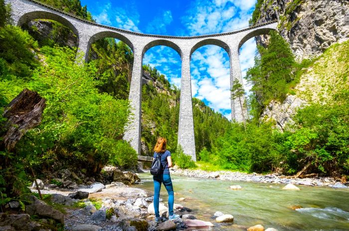 A woman gazes upward from the edge of a river, admiring a multi-arched viaduct above her.