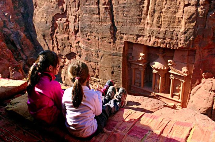 Two young girls perch on a cliff, gazing down into a valley where the ancient city of Petra is intricately carved into the red rock.