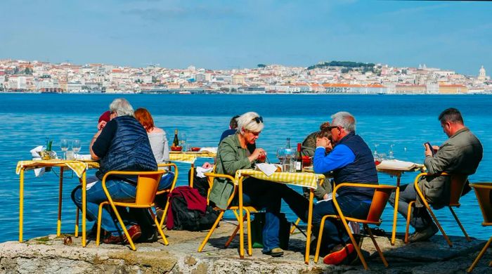 Diners enjoying meals at yellow tables along the Tagus River, with Lisbon's skyline in the background.