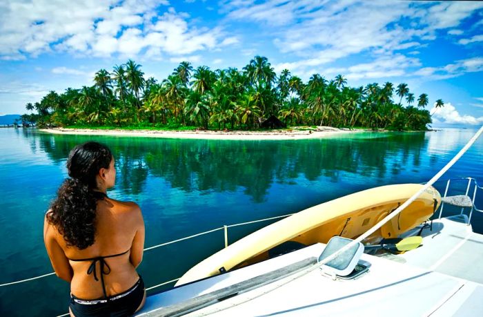 A woman sits on the edge of a boat, gazing at a picturesque island.