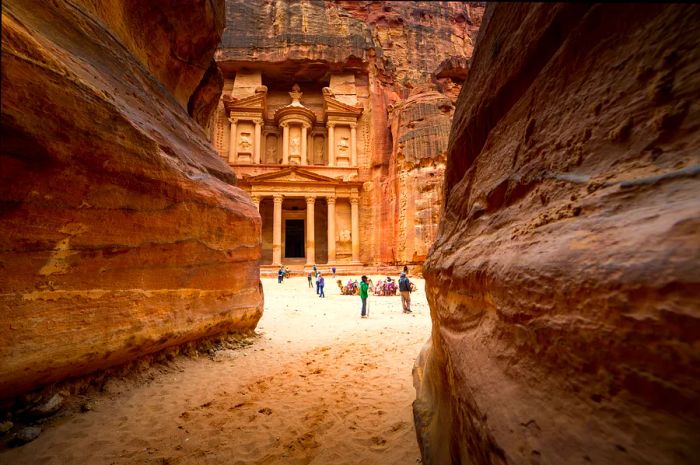 Visitors at Al-Khazneh (The Treasury) in Petra, visible at the end of a sandy trail through rocky terrain