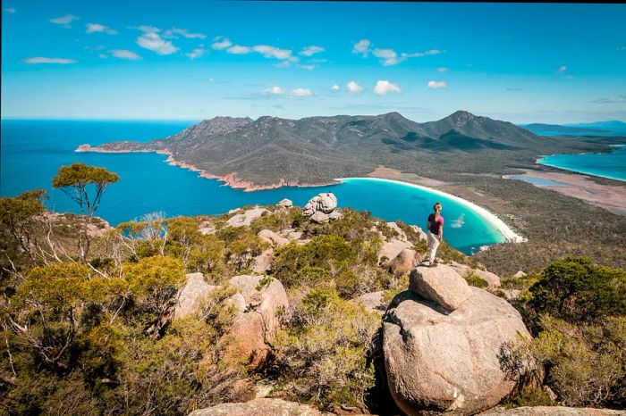 A hiker stands at a viewpoint gazing down at a stunningly curved beach.