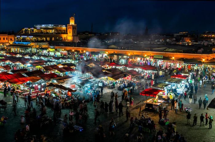 A bustling market square illuminated at night, filled with people enjoying freshly cooked dishes from food stalls.