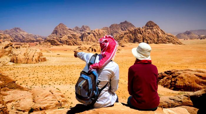 A tourist and a local Bedouin guide relax on a rock in Wadi Rum, taking in the breathtaking views from Jebel Burdah.