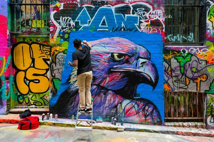 A street artist adds the final touches to a mural of an eagle on a graffiti-covered alley wall.